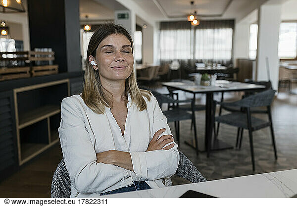 Smiling businesswoman with arms crossed listening music through wireless in-ear headphones at restaurant