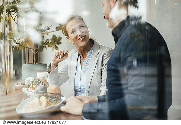 Smiling businesswoman with businessman having lunch in cafe seen through glass