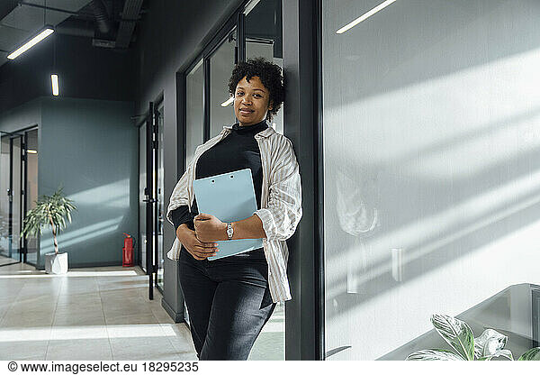 Smiling businesswoman with clipboard leaning on glass in office