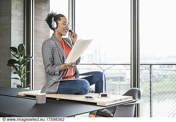 Smiling businesswoman with document talking through headset at work place