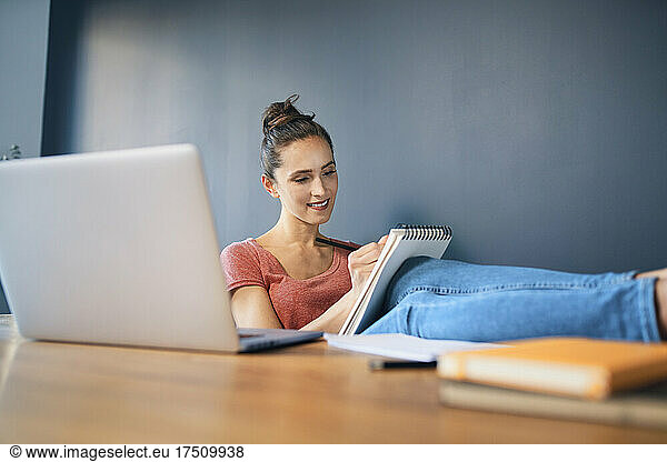 Smiling businesswoman with feet up on desk writing in note pad at home office
