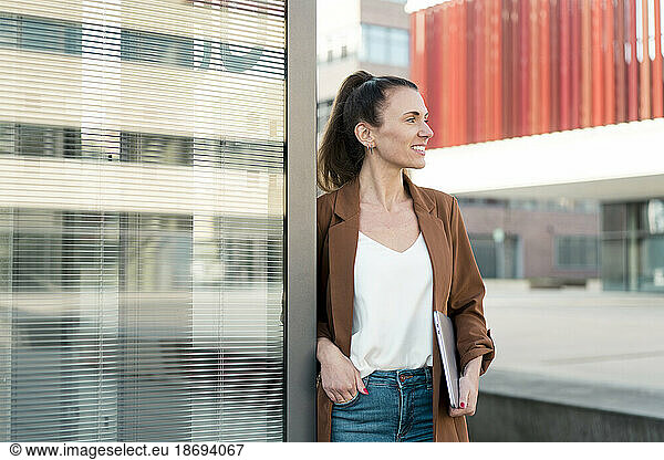 Smiling businesswoman with hand in pocket leaning on wall