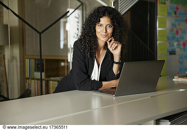 Smiling businesswoman with hand on chin leaning by laptop at workplace