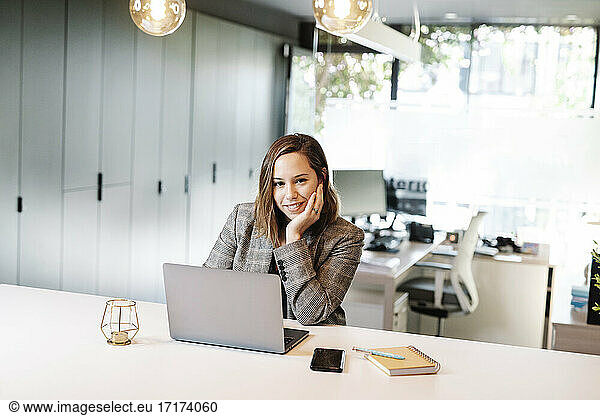 Smiling businesswoman with hand on chin using laptop at desk in office