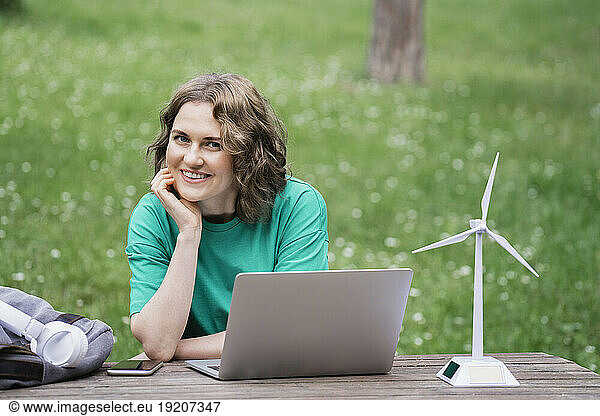 Smiling businesswoman with laptop and wind turbine model on table in nature