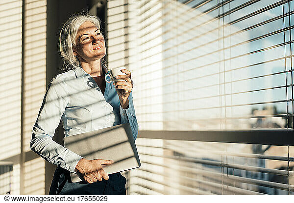 Smiling businesswoman with laptop holding coffee cup looking through window in office