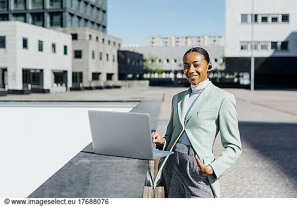 Smiling businesswoman with laptop in downtown district on sunny day