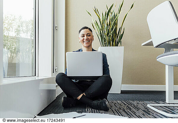 Smiling businesswoman with laptop on lap sitting on floor in office