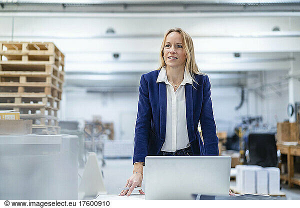 Smiling businesswoman with laptop standing at desk in factory