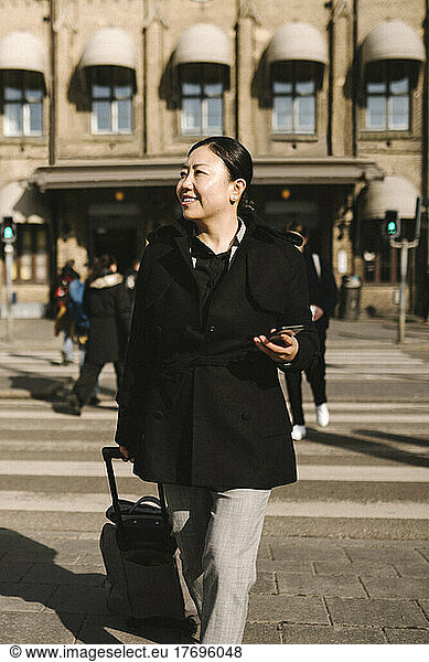 Smiling businesswoman with luggage holding smart phone while crossing street in city