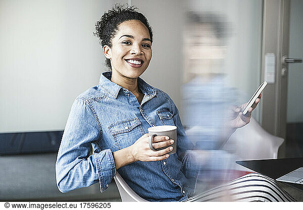 Smiling businesswoman with smart phone and coffee cup at work place