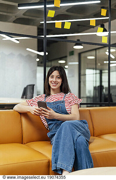 Smiling businesswoman with smart phone sitting on sofa at office