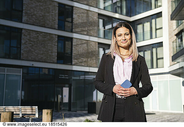 Smiling businesswoman with smart phone standing in front of office building