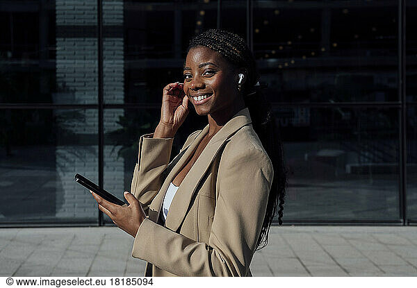 Smiling businesswoman with smart phone wearing wireless in-ear headphones at footpath