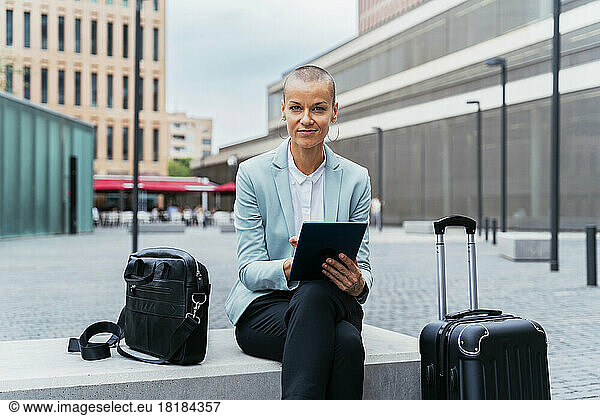 Smiling businesswoman with tablet PC and luggage sitting on bench at footpath