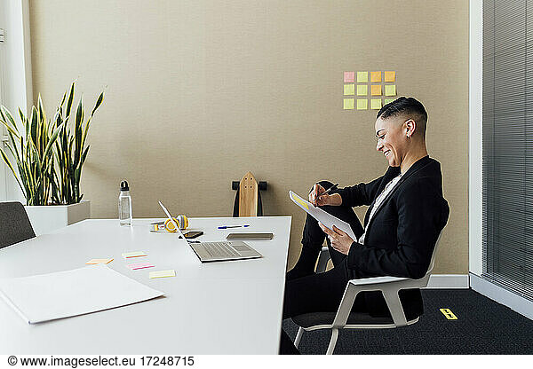 Smiling businesswoman writing on paper while sitting at desk in office