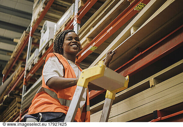 Smiling carpenter standing on ladder by rack while working in warehouse
