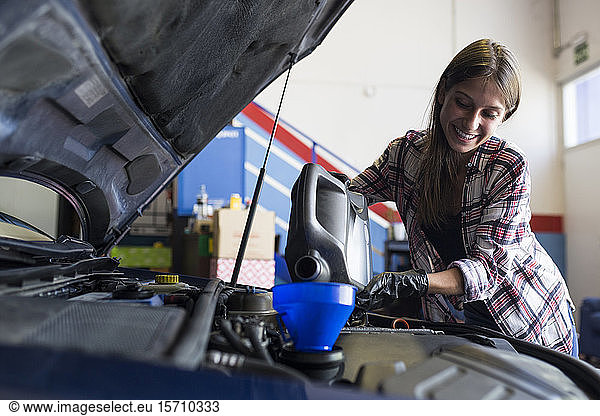 Smiling casual woman in gloves holding can of engine oil and pouring it through funnel into car working in service