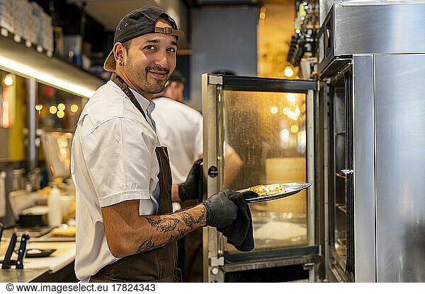 Smiling chef holding food in plate standing by oven at restaurant