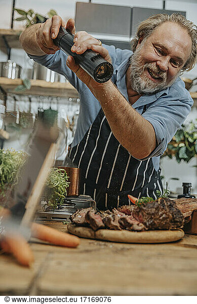 Smiling chef seasoning pepper on grilled tomahawk steak while standing in kitchen