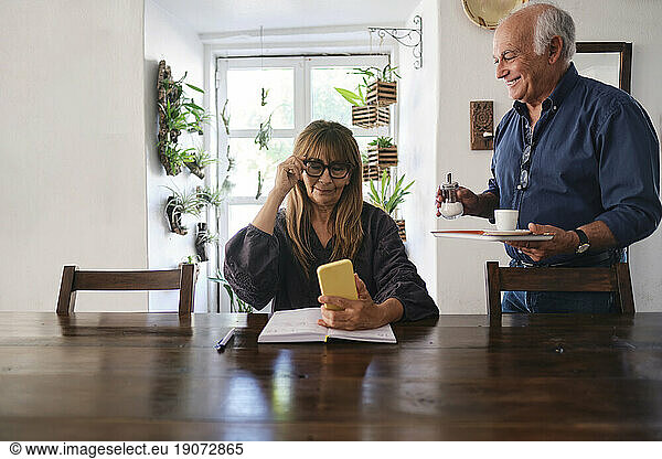 Smiling coffee shop owner serving coffee to colleague sitting with smart phone and diary at table