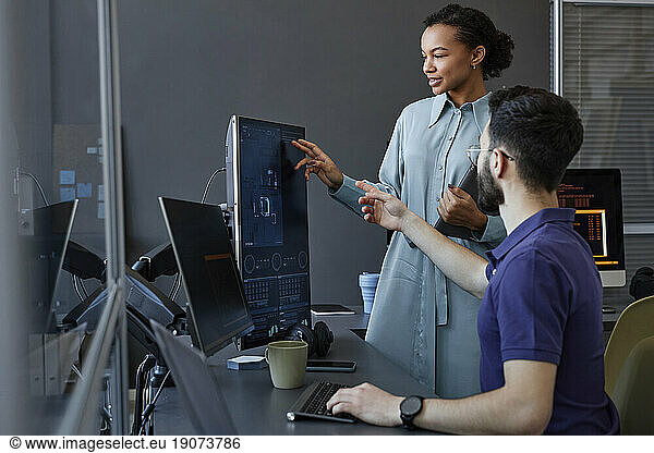 Smiling computer programmer gesturing and explaining to colleague over desktop PC