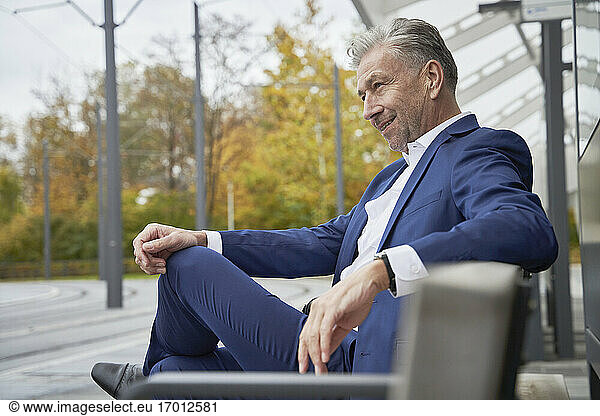 Smiling confident senior male professional sitting on bench while waiting at bus stop