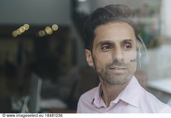 Smiling contemplative businessman wearing headset seen through glass