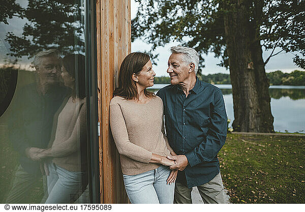 Smiling couple holding hands looking at each other by glass wall in garden