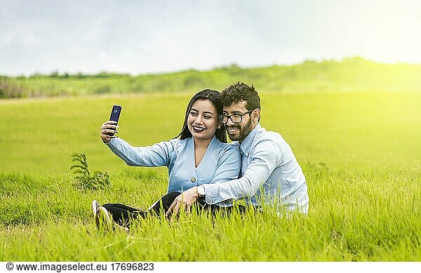 Smiling couple in love sitting on the grass taking selfies  Young couple in love taking a selfie in the field  People in love taking selfies in the field with their smartphone