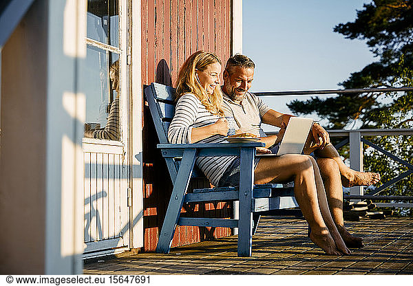 Smiling couple using laptop while having breakfast at porch in log cabin