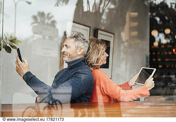 Smiling couple using wireless technologies sitting cafe seen through glass