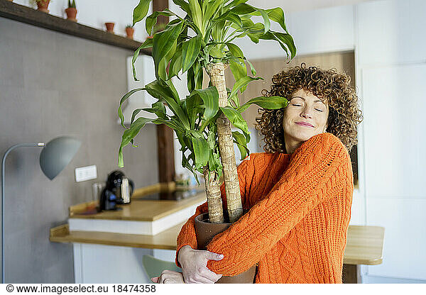 Smiling curly haired woman embracing potted plant at home
