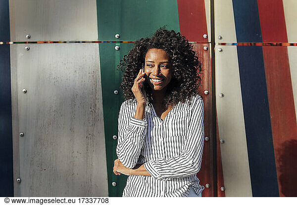 Smiling curly haired woman talking on smart phone while standing in front of wall looking away