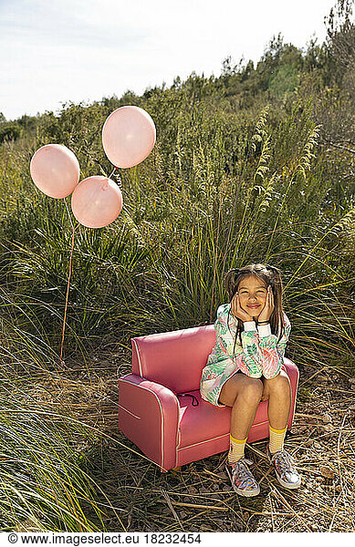Smiling cute girl sitting with head in hands on pink armchair