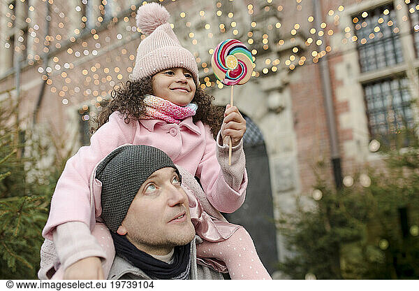 Smiling daughter holding lollipop candy and sitting on father's shoulders