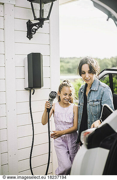 Smiling daughter with mother charging electric car outside house