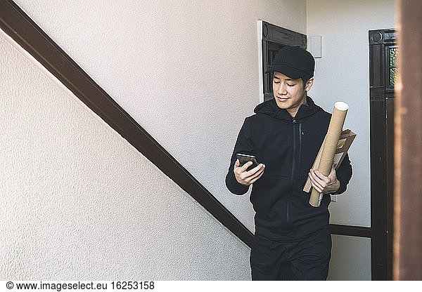 Smiling delivery man with package using phone against wall