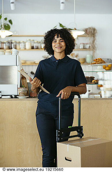 Smiling delivery woman holding clipboard and standing near package in cafe