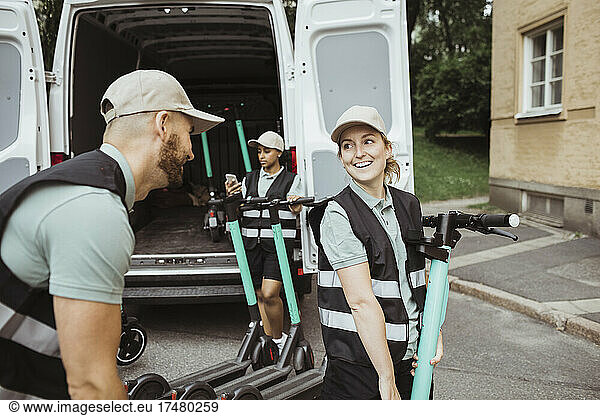 Smiling delivery woman looking at male coworker while loading push scooter in van