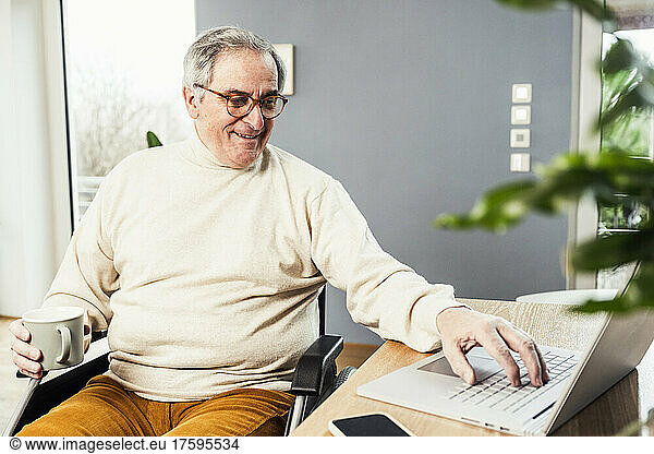 Smiling disabled man holding coffee cup using laptop sitting on wheelchair at table