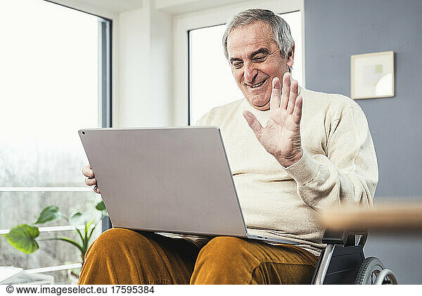 Smiling disabled man on video call through laptop waving hand sitting on wheelchair
