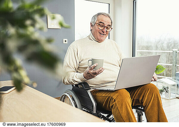 Smiling disabled senior man holding coffee cup talking on video call through laptop at home