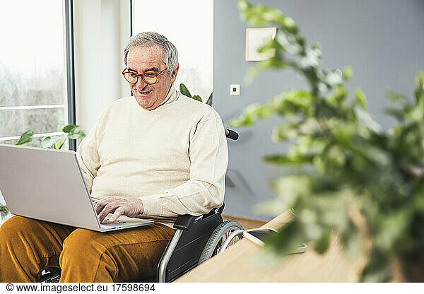 Smiling disabled senior man wearing eyeglasses using laptop on wheelchair at home