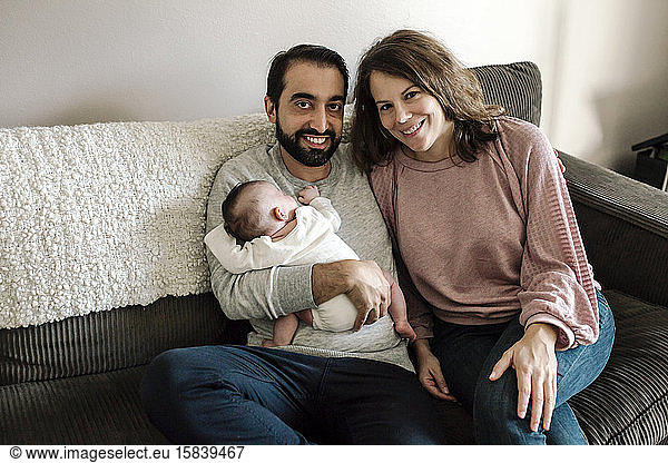 Smiling early-30â€™s parents on sofa holding newborn baby