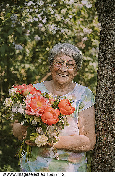 Smiling elderly woman holding fresh peony bouquet by tree trunk at garden