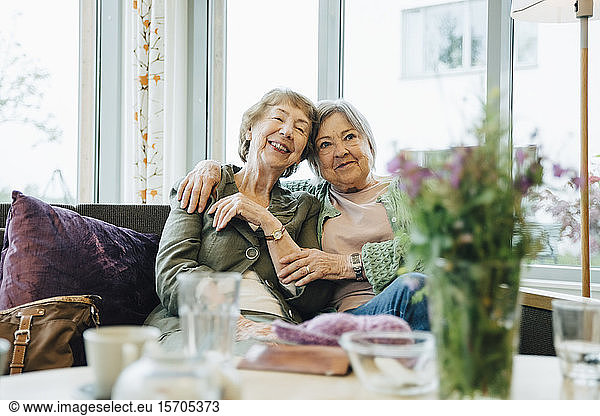 Smiling elderly women sitting with arm around on sofa against window at retirement home
