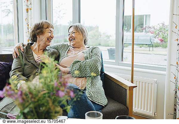 Smiling elderly women sitting with arm around while looking at each other on sofa against window at retirement home