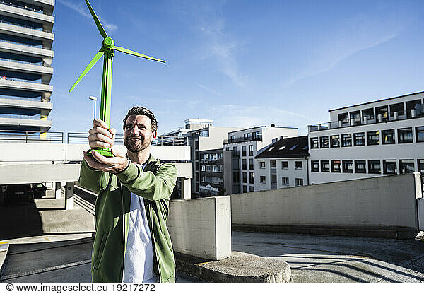 Smiling engineer holding green wind turbine model at terrace on sunny day