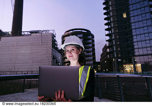 Smiling engineer holding laptop in front of building at dusk